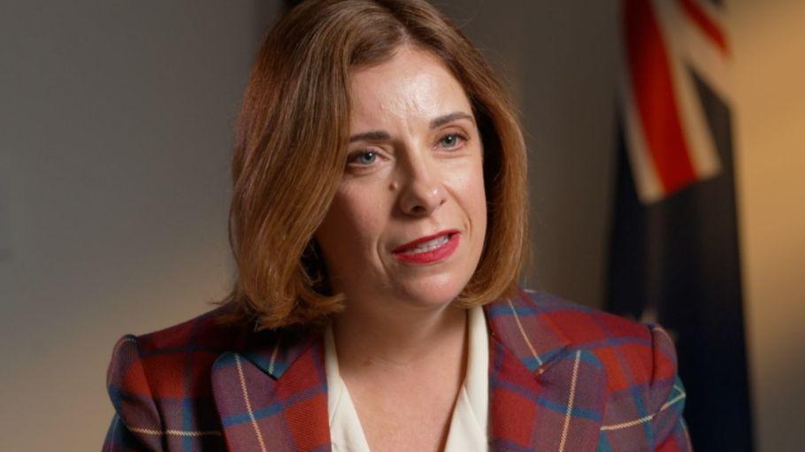 A woman in a tartan jacket and white dress speaks. Several flags can be seen in the background