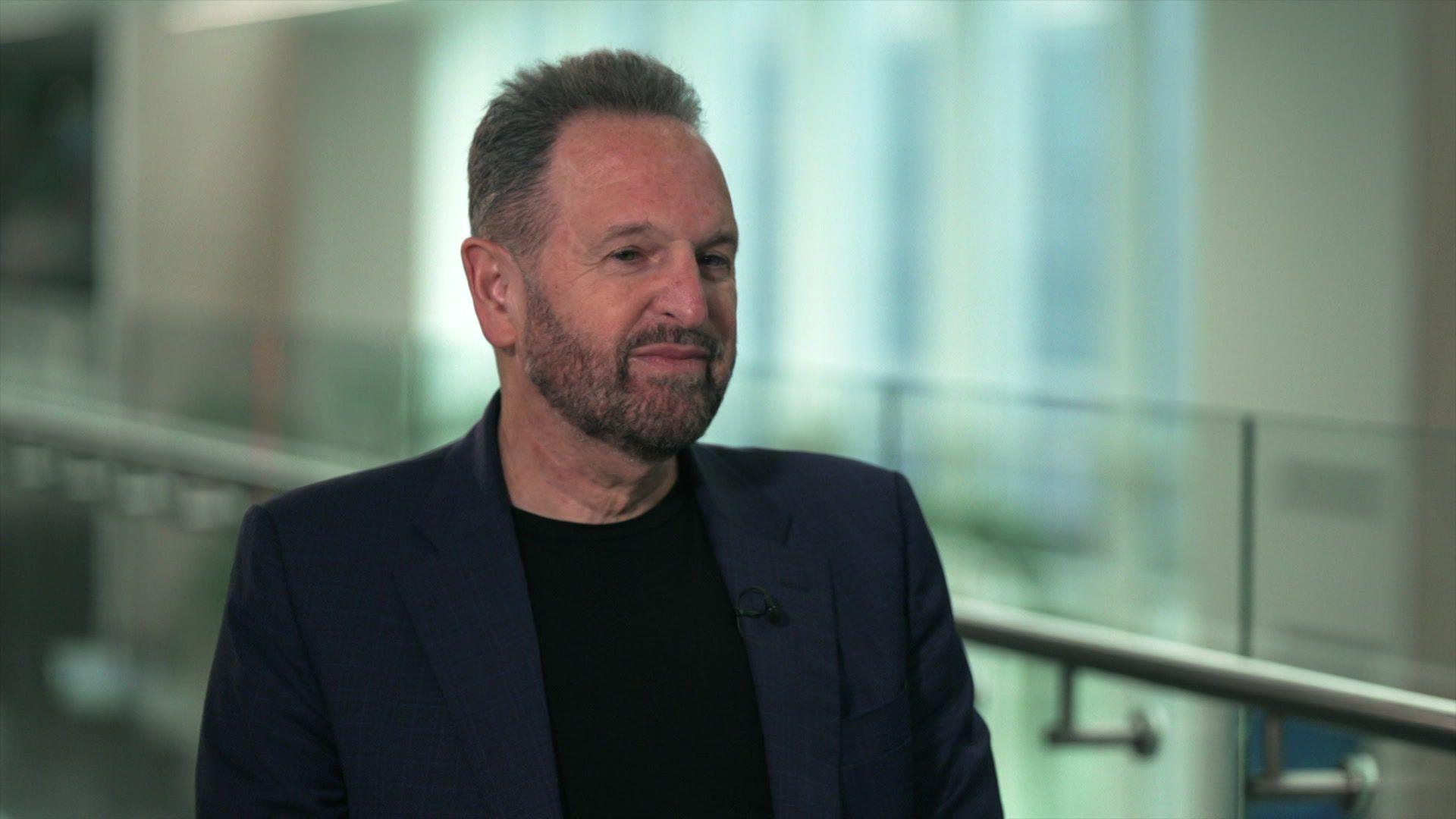 Rene Haas sitting being interviewed. He has short brown hair and a short beard, and is wearing a dark blue blazer over a black T-shirt.